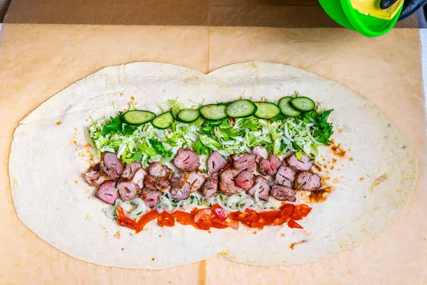 Dilimlenmiş etler, salatalık, salatalık ve domateslerle tortilla üzerine sarılmış bir paket hazırlanıyor. Malzemelerin canlı renkleri iştah açıcı bir görüntü oluşturuyor..