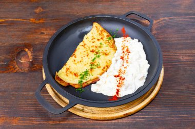 Lezzetli bir omlet dökme demir bir tavada sunulur, yanında bir parça kremalı yoğurt sosu ve baharatlarla serpiştirilir. Sahne, ahşap bir masa ayarını yakalıyor..