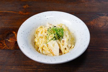 Kremalı bir makarna tabağı zarif bir şekilde beyaz bir kasede sunulur, üzeri de narin yeşilliklerle kaplıdır. Kırsal ahşap masa yemek deneyimine sıcaklık katar ve onu davetkar kılar..