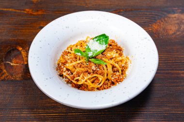 Spagetti, rendelenmiş peynir ve taze fesleğenle kaplanmış kırsal beyaz bir kaseyle kaplıdır ve koyu ahşap bir arka plana karşı iştah açıcı bir sunum yaratır..