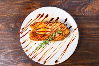 Güzel bir şekilde sunulmuş ızgara somon filetosu beyaz bir tabakta oturur. Yaratıcı bir sos çiselemesi ve taze otlarla zenginleştirilir..