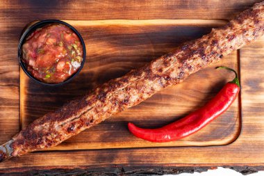 Uzun, ızgara bir kebap, ahşap bir tahtanın üzerinde bir kase taze salsa ve kırmızı acı biberin yanında oturur. Parlak ışık, yemeğin leziz dokusunu ve rengini vurgular..