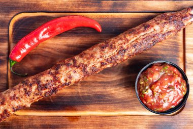 Uzun bir kebap mükemmel bir şekilde ızgara edilir ve bir kase taze salsa ve canlı kırmızı biberin yanına ahşap bir tabağa yerleştirilir. Yemek zengin lezzetler ve dokular sergiliyor..