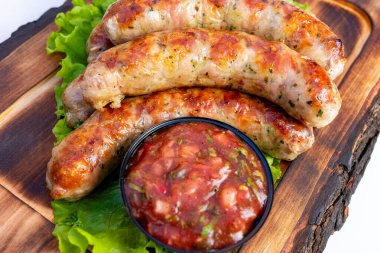 Sulu ızgara sosisler taze yeşil marulun yanında ahşap bir tabakta düzenlenir. Bir kase baharatlı salsa tat katar, canlı bir mutfak sunumu sunar..