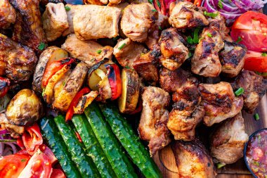 Canlı bir ızgara et ve ahşap tabakta sıralanmış renkli sebzeler. Taze malzemeler arasında biber, salatalık ve domates yer alıyor..