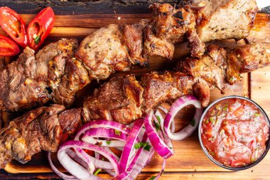 Dilimlenmiş soğan ve taze acı biberle birlikte lezzetli ızgara et şişleri ahşap bir tahtanın üzerinde sunuluyor. Küçük bir kase zengin sos tabağa baharatlı bir dokunuş ekler..