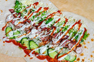 Renkli bir ambalajda ızgara tavuk, kıtır marul, salatalık ve üzerine sanatsal olarak serpiştirilmiş çeşitli soslar yer alıyor. Bu taze sokak yemekleri gündelik yemek ortamında canlı bir tat deneyimi sunuyor..