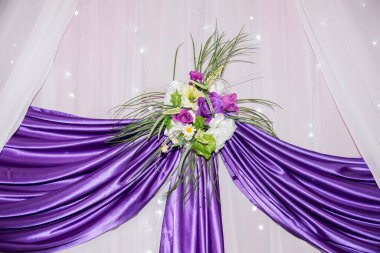 Çiçek ve yeşillik karışımıyla süslenmiş güzel bir şekilde düzenlenmiş mor kumaş her olay için çarpıcı bir zemin oluşturur. Yumuşak ışıklandırma ayarların zarafetini arttırır.