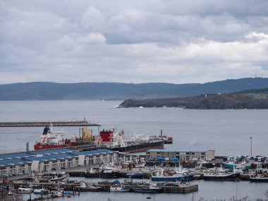 Bir Corua, İspanya; 03-16-24: Arkasında dağlar ve kayalıklar bulunan yağmurlu ve bulutlu bir günde A Corua limanına yanaşan yük gemileri ve balıkçı teknelerinin görüntüsü