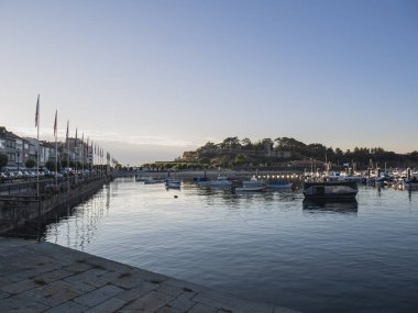 Baiona, Pontevedra, İspanya; 07-27-2024: Baiona limanına yanaşan teknelerin görüntüsü, su boyunca bayrak örtülü gezinti güvertesi ve gün batımında tarihi Monterreal Kalesi