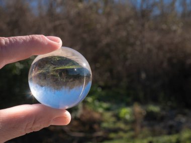 Two fingers hold a small transparent glass ball that reflects the vegetation of the trees, branches and the sky but in reverse in the ball itself. clipart