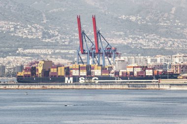 Malaga, İspanya; 07-06-2025: Konteyner gemisi MSC Dominique büyük vinçler ve renkli konteynırlarla ticari bir limana yanaştı. Arka planda sahil kenti ve tepeler