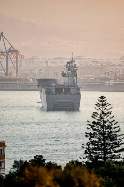 Malaga, İspanya; 07 / 07 / 2025: Gün batımında işlek limandaki askeri gemi ve konteyner gemisi, arka planda dev vinçler ve şehir silüeti bulunuyor
