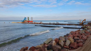 İnşaat makineleri, fırtınalı Baltık deniz kıyısında kıyı koruma çalışmalarını güneşli kış günlerinde büyük kayalar döşeyerek ve kazık çakarak yürütüyor. İnsansız deniz manzarası.