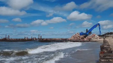 İnşaat makineleri kepçesi, yaz günü büyük kayalar döşeyerek bulutlu Baltık deniz kıyısında kıyı koruma çalışmaları yürütüyor. Yaz mevsimi deniz manzarası.