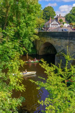 Turistler iki sandalda oturup kemer köprüsüne doğru kürek çektiler. Etrafı yeşil ağaçlarla çevrili, arka planda Dünya 'nın Birahanesi, Knaresborough, İngiltere.
