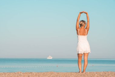 Deniz kıyısında beyaz elbiseli bir kız ellerini kaldırıyor.