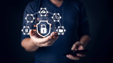 Karanlık bir ortamda veri koruması, güvenli ağlar ve teknoloji koruyucularını temsil eden birbirine bağlı dijital simgelerle çevrili siber güvenliği simgeleyen parlayan bir kalkan simgesi tutan bir kişi..