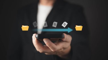 Bir kişi, sarı klasörler arasında dijital belge transferini gösteren, kusursuz dosya paylaşımını, belge yönetimini ve verimli dijital organizasyonu sembolize eden bir akıllı telefon tutuyor.