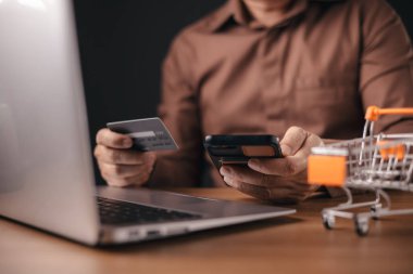 Kahverengi gömlekli bir adam bir dizüstü bilgisayar üzerinde çalışırken kredi kartı ve akıllı telefon kullanarak online ödeme yapıyor. Küçük bir alışveriş arabası dijital alışveriş, e-ticaret ve mali işlemleri sembolize eder.