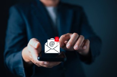 Takım elbiseli işadamı, akıllı telefonunun üzerinde gezinen dijital bir e-posta bildirim simgesi ile etkileşime geçiyor. Sembollü ve kırmızı alarmlı parlayan zarf posta, gelen kutusu güncellemeleri ve iletişimi simgeliyor.