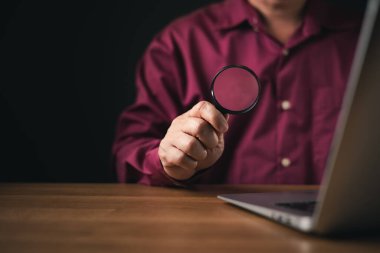 Bir adam dizüstü bilgisayarının yanında bir büyüteç tutarak soruşturmayı, araştırmayı veya incelemeyi sembolize eder. Koyu arkaplan analizi, siber güvenliği, denetimi ve dijital güvenliği vurguluyor.