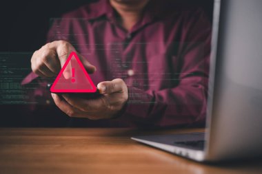 Bir adam elinde kırmızı alarm sembolü olan bir akıllı telefon tutuyor. Siber güvenlik tehditlerini, veri ihlallerini, hacklemeyi ya da kötü amaçlı yazılımı temsil ediyor. Dijital arkaplanda güvenlik riskleri vurgulanıyor.