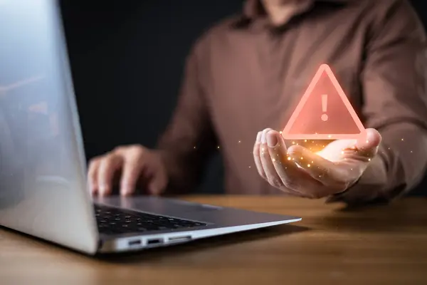 Dizüstü bilgisayar kullanan işadamı parlak bir uyarı simgesi, sembol uyarısı ve siber güvenlik tehdidi sunuyor. Fütürist dijital alarm potansiyel riskleri, veri ihlallerini ve çevrimiçi güvenlik kaygılarını vurguluyor..
