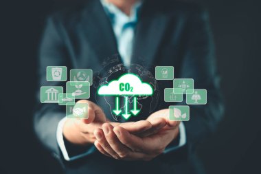 İş adamı, yeşil sürdürülebilirlik ikonu ile çevrili karbondioksit azaltımının parlayan dijital görüntüsünü tutuyor. Görüntü çevresel sorumluluğu, karbon ayak izini azaltmayı ve çevre dostu girişimleri sembolize ediyor.