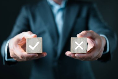 A businessman in a suit presents two wooden blocks, one with a white checkmark and the other with a white X, symbolizing decision making, evaluation, approval, rejection, and business choices.