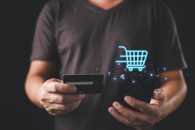 Bir kişi kredi kartı ve akıllı telefon tutarken, yukarıda parlayan dijital bir alışveriş arabası simgesi, mobil ticareti, çevrimiçi alışverişi ve dijital ödeme işlemlerini sembolize ediyor.