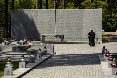 Catholic priest stands near Memorial wall with the names of Poles, victims of Stalinist repressions at Polish military cemetery Polish graves Bykivnia, near Kyiv, Ukraine 2024, May 19 clipart