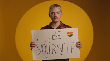 Pozitif genç gay adam elinde Igbt sloganıyla fotoğraf makinesine gülümsüyor.