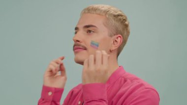 Yanaklarında gökkuşağı bayrağı olan, pembe gömlekli, kameraya gülümseyen eşcinsel bir adam.