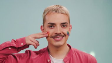 Stüdyoda yanaklara gey gökkuşağı bayrağı çizen mutlu sarışın adamın ön çekimi.