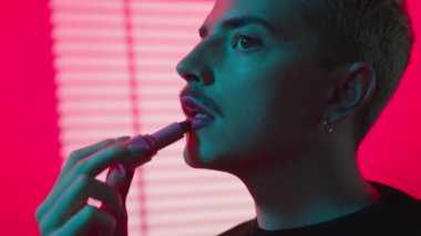 Genç, sarışın, bıyıklı, neon pembe stüdyosunda ruj süren eşcinsel bir adam.