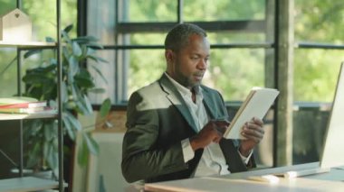 Yakından bakınca sakallı, yakışıklı Afro-Amerikan bir adam ofis teknolojisi işadamında gün ışığında kameraya bakarak tablet bilgisayar kullanıyor. İşçi. Yavaş çekim