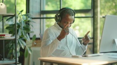 Mutlu, başarılı, başarılı, Afro-Amerikan erkek ofis çalışanı mutlu gülümseyen bir bilgisayar ve kulaklık kullanıyor. Ofiste sandalyede dans etmek. Yavaş çekim