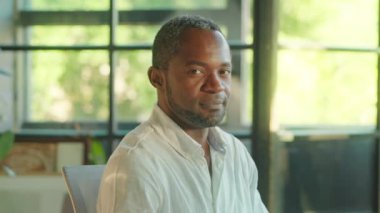 Mutlu bir Afrikalı Amerikalının portresi gülümseyerek kameraya bakmak için başını çeviriyor. Ofiste oturuyor. İş adamı, girişimci, CEO. Şirket işi. Yavaş çekim