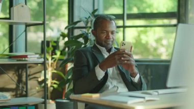 Çekici bir Afrikalı Amerikalının portresi internet üzerinden akıllı telefon mesajlaşması kullanıyor. İş yerindeki modern güneş ışığı ofisinde oturmuş gülümsüyorum. Modern aygıtlar, işyeri
