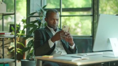 Yakışıklı Afro-Amerikan adam internet üzerinden iletişim kurmak için akıllı telefon mesajları kullanıyor. Masada ciddi bir şekilde oturup modern güneş ışığı ofisinde gülümsüyorsun. Modern aygıtlar, işyeri
