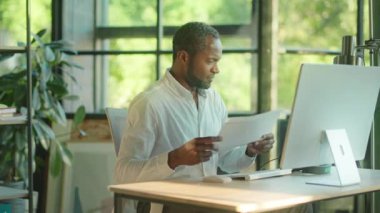 Yakın çekim başarılı genç Afro-Amerikan ofis çalışanı elinde bilgisayar ve kulaklıkla gülümseyen kağıtlar tutuyor. Yavaş çekim