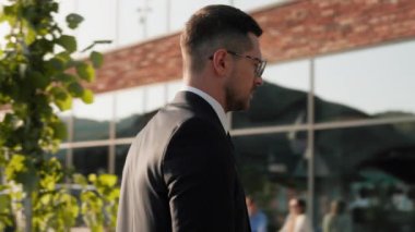 Takım elbiseli, modern binanın yanında duran endişeli iş adamı gün ışığından rahatsız oluyor. Girişimci adam. Kamera hareket ediyor