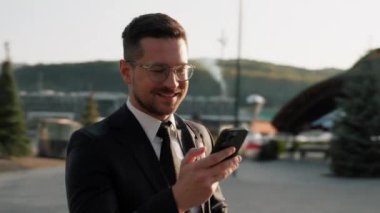 Takım elbiseli ve gözlüklü bir portre işadamı elinde akıllı bir telefonla modern binanın yanında durup etrafına bakıp gün ışığına gülümsüyor. Erkek girişimci.