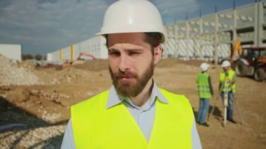 Kameraya bakan kendinden emin beyaz erkek mühendisin görüntüsü. Yüksek görüşlü yelekli ve koruyucu kasklı bir adam. Güneşli bir gün. İnşaat alanı. Açık havada.