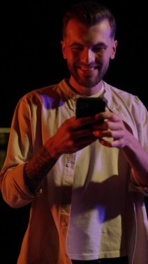 Gülümseyen adamın akıllı telefona bakıp sohbet edişinin görüntüsü. Telefonu kullanan güzel sakallı genç adamın portresi. Açık havada, gece. Hippi. Anamorfik lenslerle dikey çekim. Mor ışık