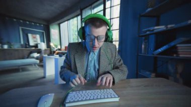 Gözlüklü ve kulaklıklı duygusal adam ekranda klavyede bilgisayar kullanıyor. İnek. Aktif iş. Ofis ya da daire