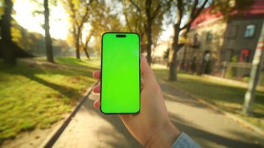 Sonbahar parkında yeşil ekran ve krom anahtarlı akıllı telefonu tutan genç bir erkeğin yakın görüntüsü. Telefon kullanan, sarı yapraklı ağaçların bulanık arka planında mesajlar okuyan bir adam.