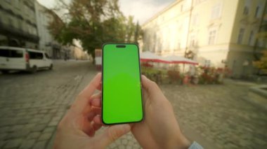 Şehir sokağının bulanık arka planında adamın ellerinin telefonu tutarken ve yeşil ekrana dokunurken yakın plan görüntüsü. Sosyal medyayı kontrol eden kişi. Krom anahtar. Mesajlaşma. Dikey pozisyonda göster. Açık havada.