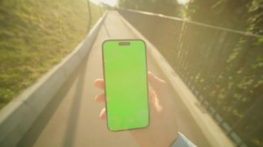 Köprüde yürürken elinde yeşil ekran olan akıllı telefonu tutan genç bir adamın görüntüsü. Dışarıda boş ekranlı cep telefonu kullanan bir adam. Doğa. Yaz zamanı.
